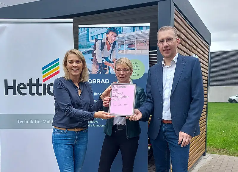 Monika Titze, von der Hettich Gruppe, steht zusammen mit einer Frau und einem Mann vor einem Poster mit dem Schriftzug Hettich und dem Hettich Logo. Sie alle tragen blaue Geschäftskleidung